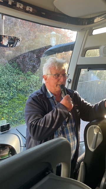 Ein grauhaariger Mann mit Brille steht im Reisebus und schaut in Richtung der Reisegesellschaft.. Er spricht in ein Mikrophon, das er in seiner rechten Hand hält.